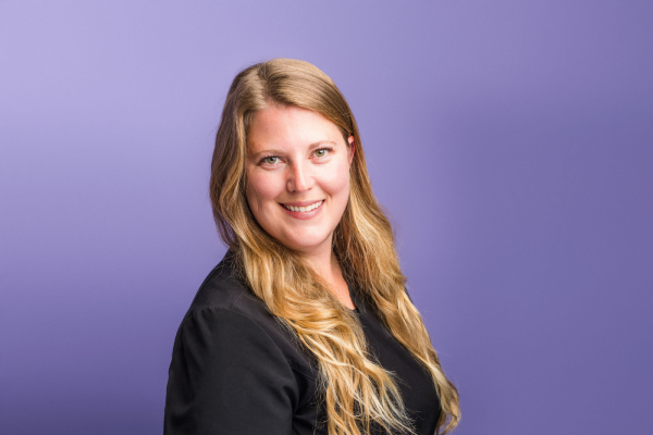 A woman with shoulder-length blonde hair is smiling at the camera. She has glasses on and is wearing a black top.